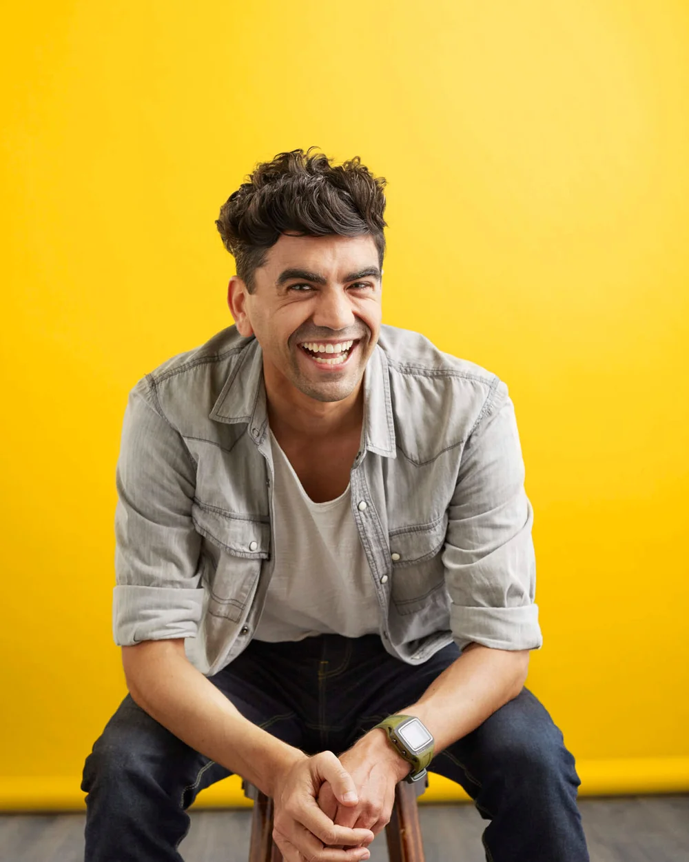 A smiling man with dark hair wearing a light denim jacket, white t-shirt, and a watch, sitting on a stool against a yellow background.