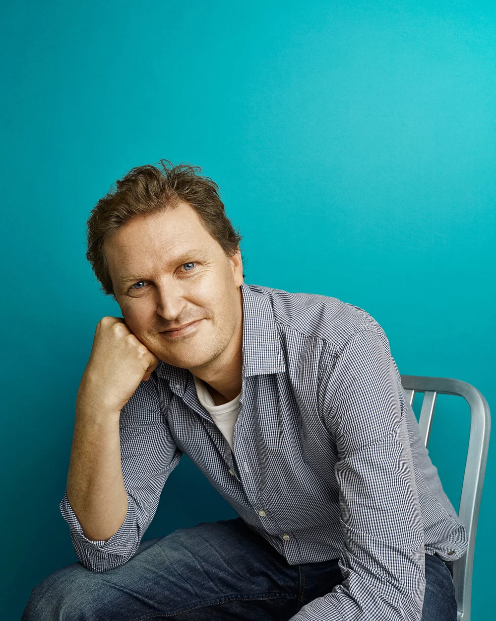 A man with brown hair and blue eyes sitting on a gray chair against a teal background, resting his head on his left hand and smiling at the camera.