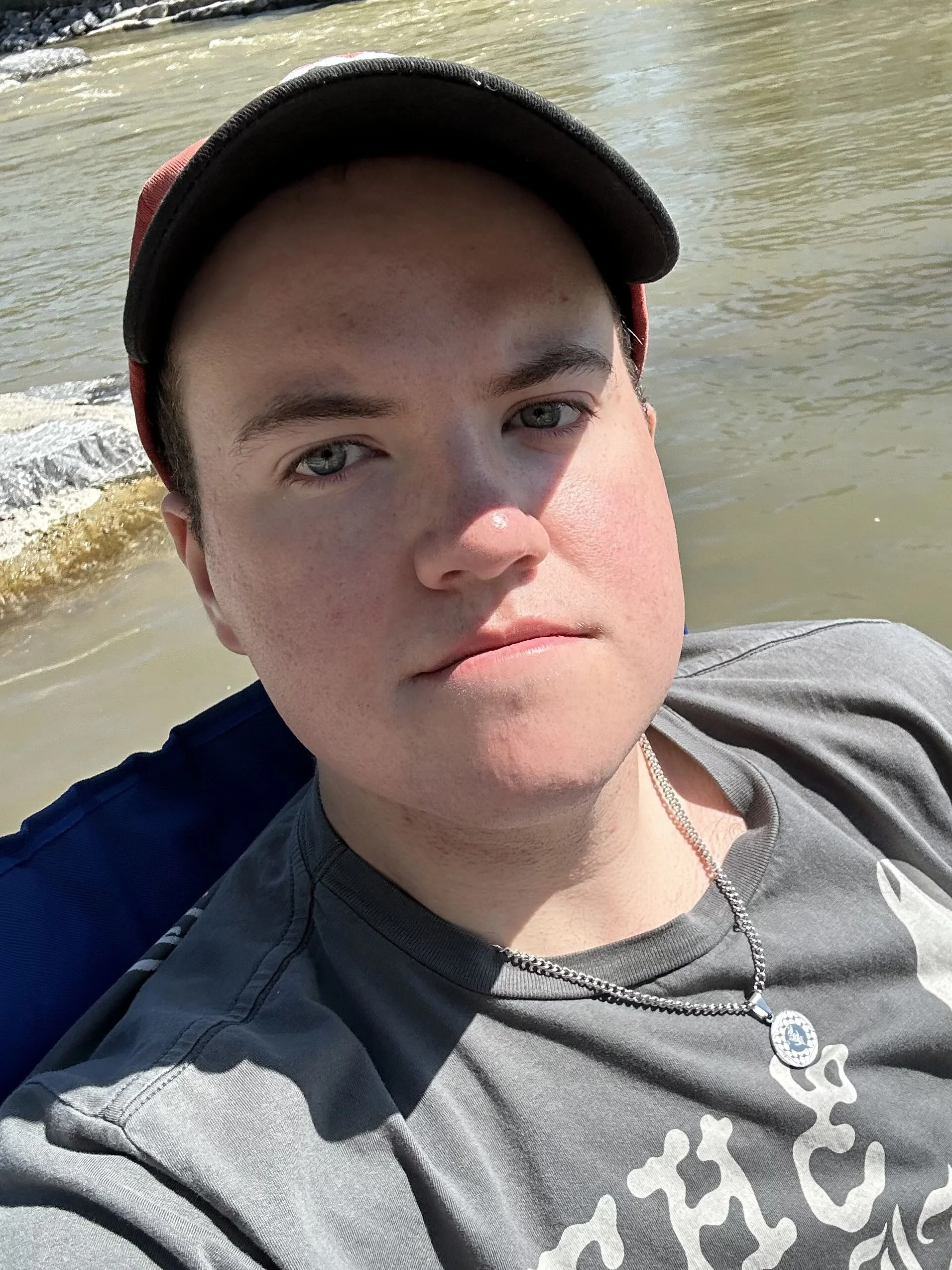 Eli Buechler is wearing a cap and a grey T-shirt with a silver chain. He is sitting outside in the sun in front of a river.