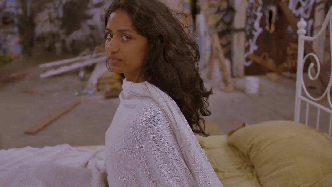 Film still of a South Asian woman with long hair, wrapped in a sheet on a bed, positioned in the middle of a decaying church covered in graffiti.
