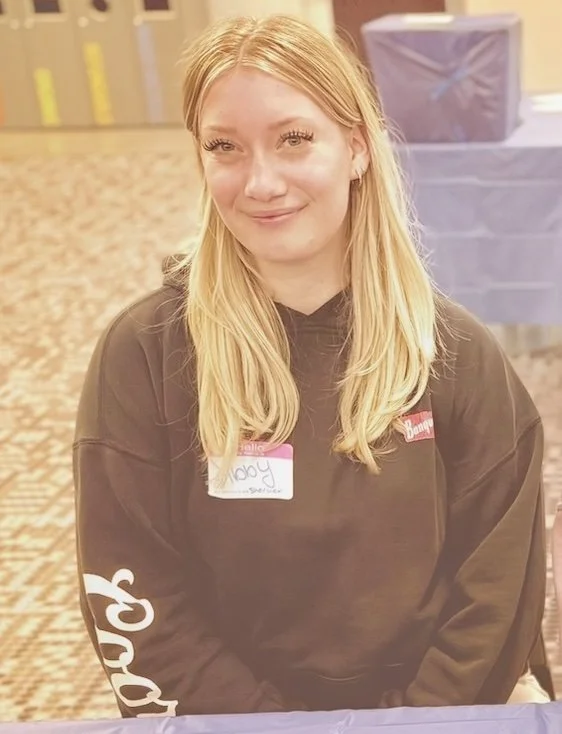 Abigail Johnson is sitting at the welcome desk at the event. She has long blond hair and is wearing a name-tag.