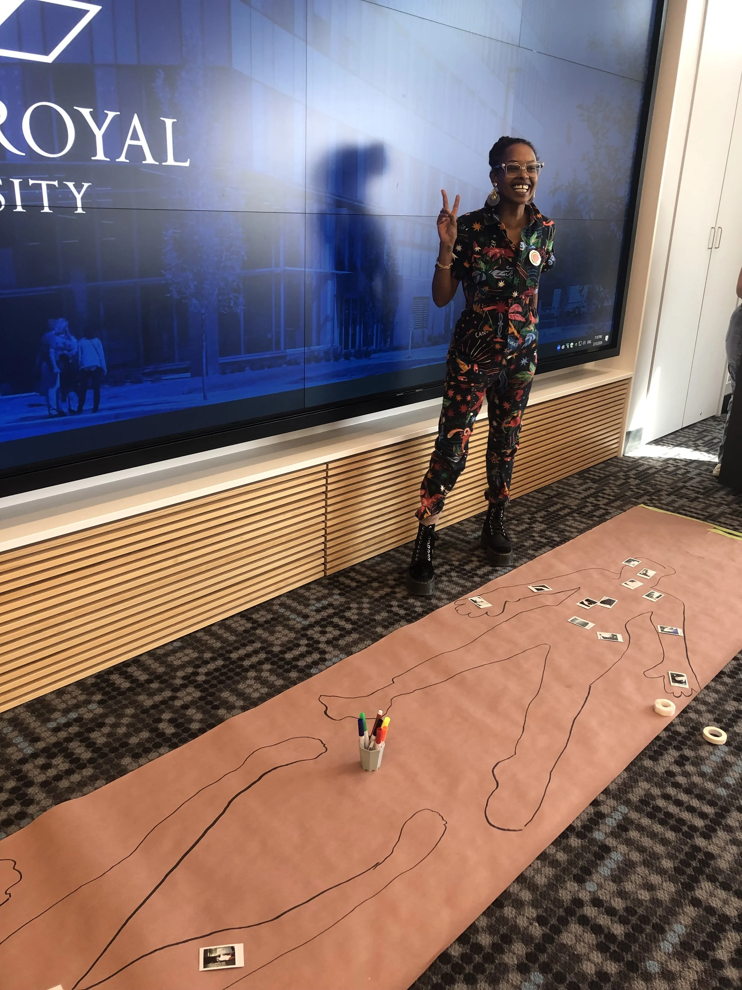 Qrescent Mali Mason poses after her 2024 keynote talk next to a tracing of her body on brown paper, populated with instant camera photos that audience members took of a part of their body where they locate thinking happening.