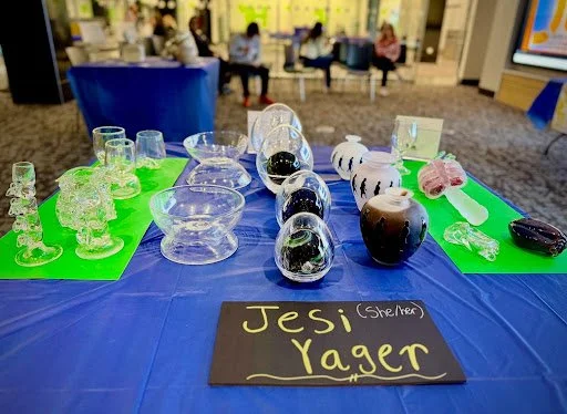 various pieces of glass displayed at a convention, with a black piece of paper in the front reading "Jesi Yager (she/her)" in yellow marker.