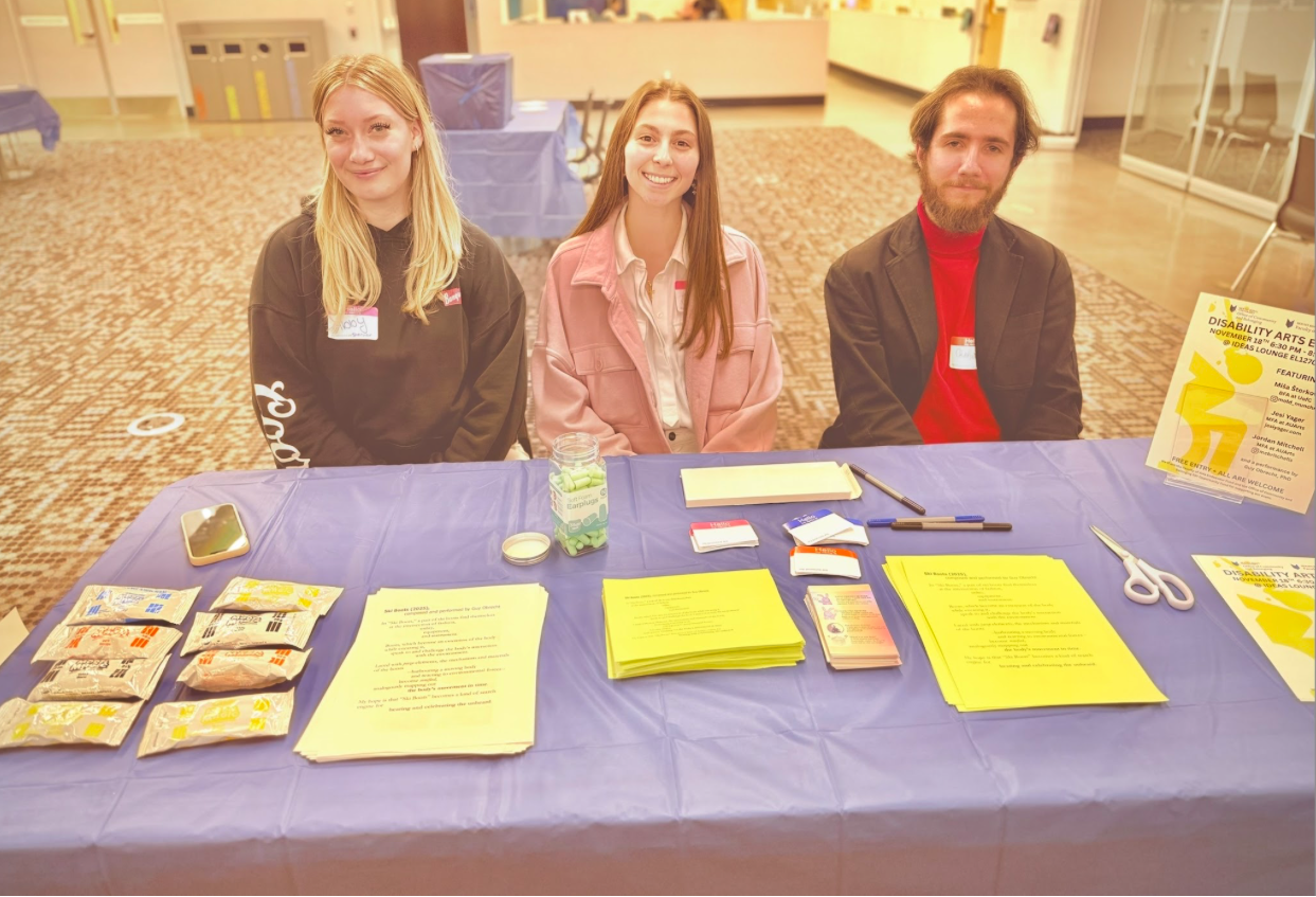 Three student co-creators sit at the welcome desk that they have set up at the pop-up event.  In front of them are an array of resources and materials that they will hand out to participants.