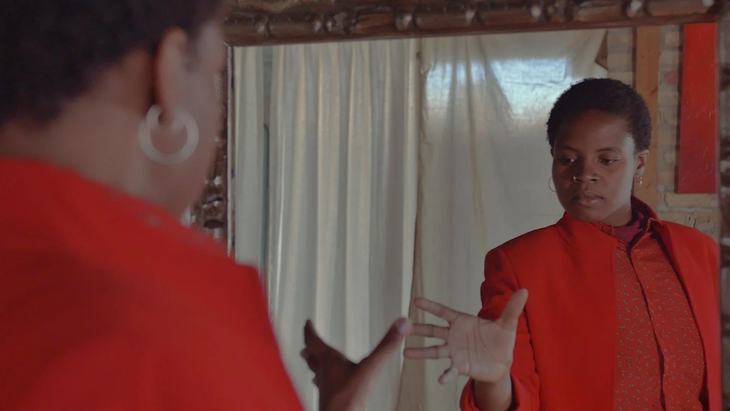 Film still of a Black femme figure wearing layers of red clothing reaches toward a mirror.