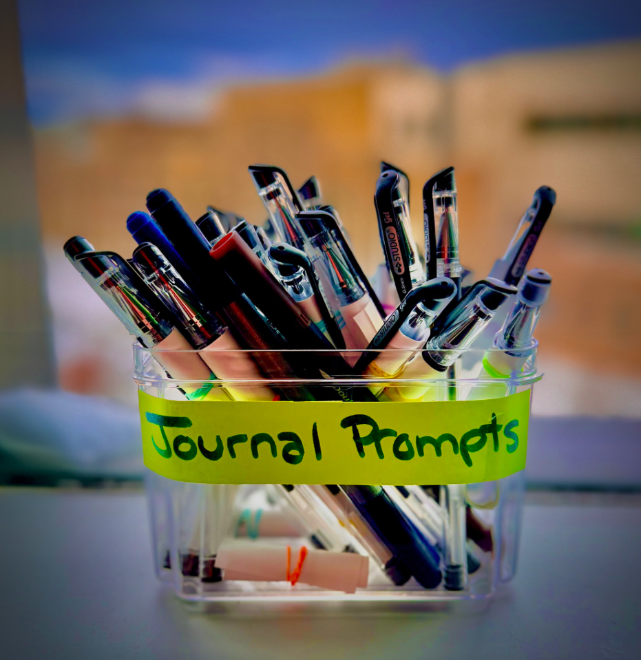 A close-up photo of pens in a container with a sign that reads journal prompts.