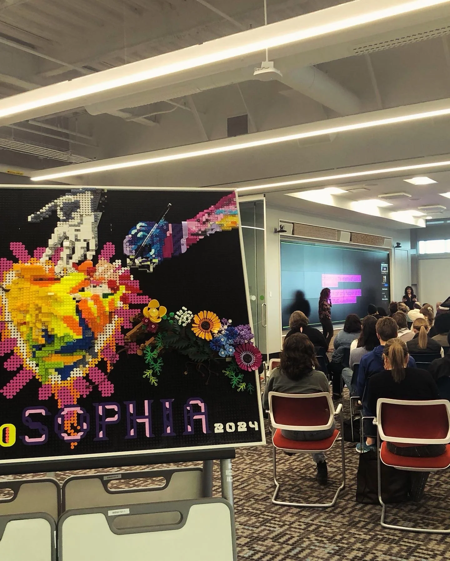 In the foreground, a LEGO bas relief by Bailey Szustak that would later become our book cover, with five hands reaching in to contribute to making a heart sculpted out of paper hands. In the background, Natalie Loveless is giving her 2024 keynote.
