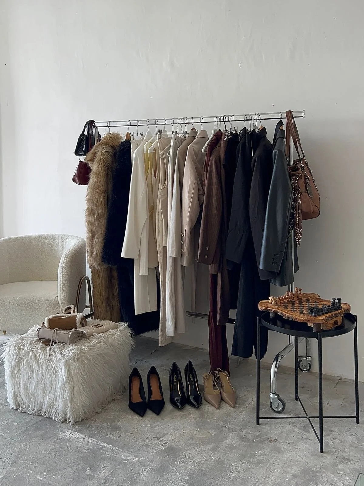 A clothing rack with various coats and jackets, a pair of heels, and a leather handbag. A small table with a chessboard and a chair with a furry covering and a handbag on it.