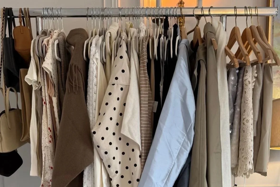 Clothing rack with various shirts, blouses, and dresses in neutral and light colors, including polka dot and striped patterns.