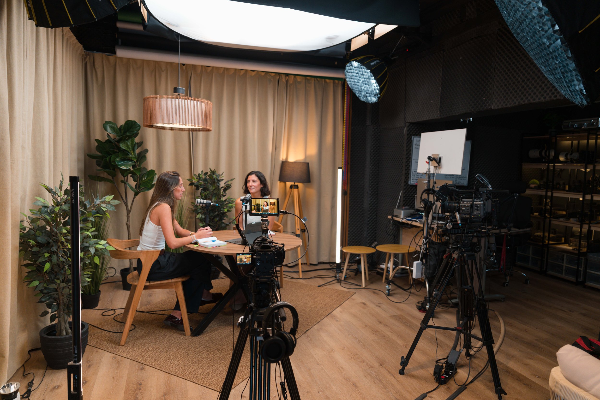 Dos mujeres grabando una entrevista en un estudio con equipo de grabación y cámaras, con cortinas beige y plantas verdes en el fondo.