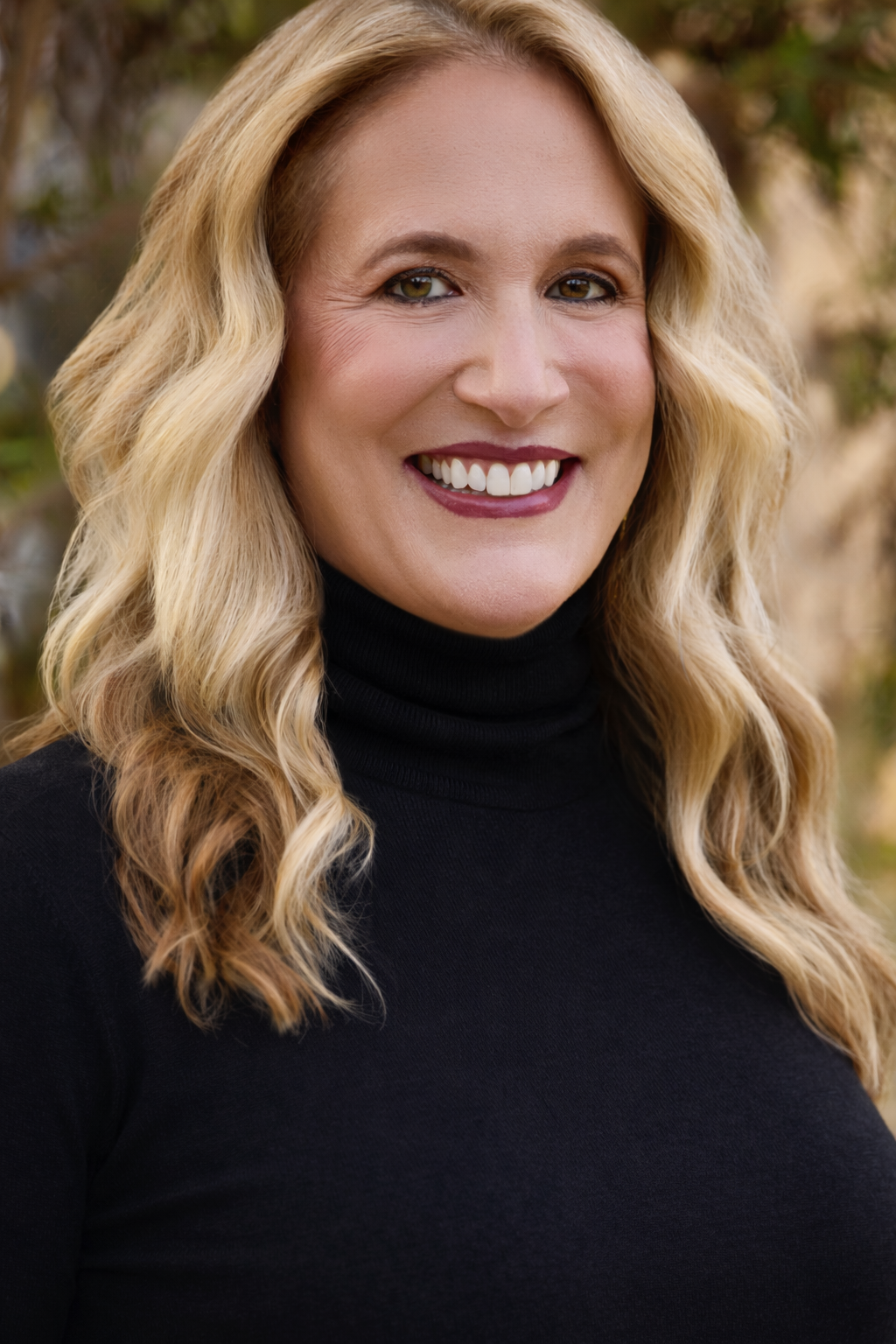 Photo of Elizabeth Elliott. A woman smiling with wavy blonde hair, wearing a black turtleneck, outdoors in a natural setting.