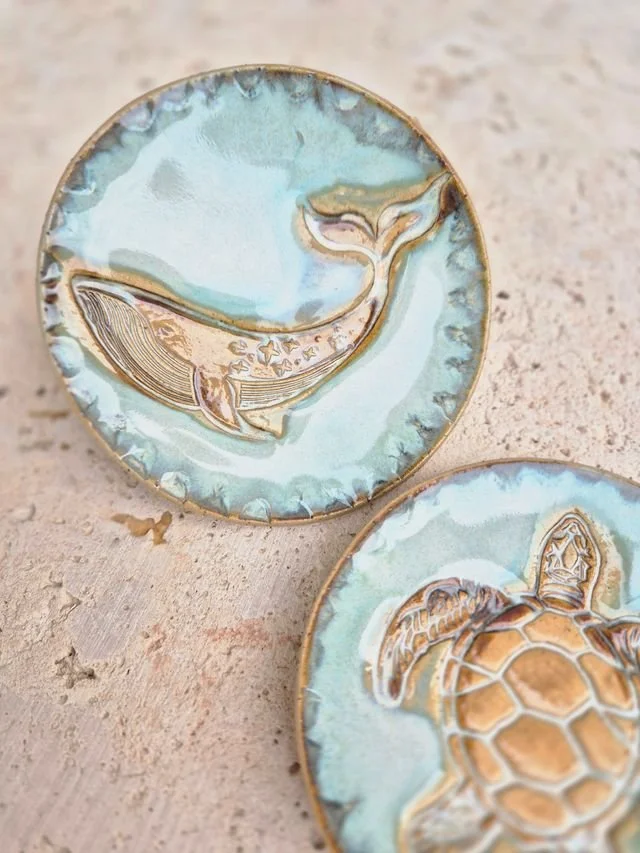 Close-up of two circular enamel pins with ocean-themed designs, one featuring a whale and the other a turtle, placed on a textured surface.