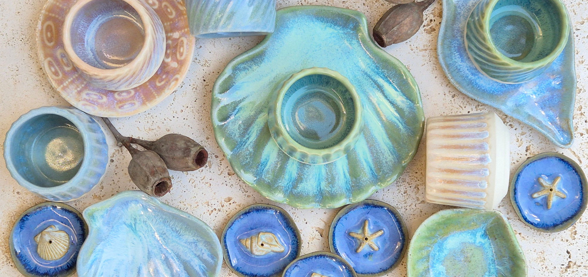 A collection of colorful ceramic shell-shaped dishes and bowls arranged on a speckled surface, with a few dried seed pods and starfish decorations among them.
