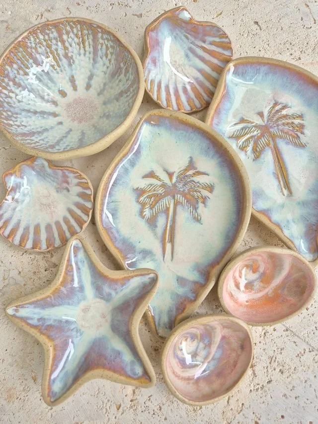 Collection of ceramic dishes with beach-themed designs, including palm trees, shells, starfish, and abstract patterns, on a textured surface.
