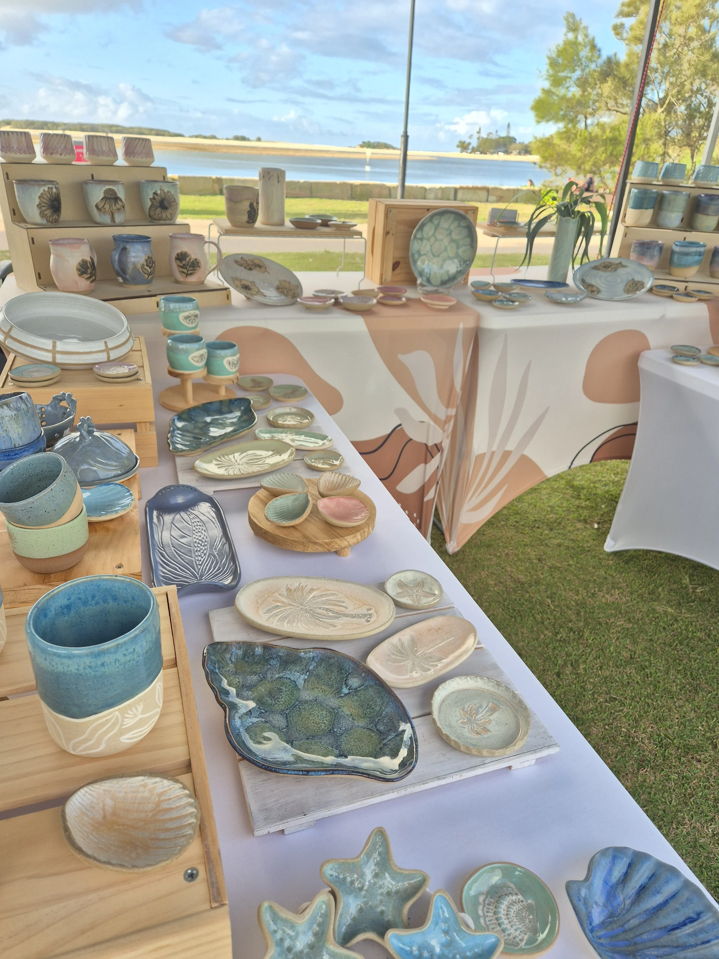 A wider view of a table set outdoors in a park, displaying a curated collection of handcrafted ceramics from the store, including plates, cups, and a vase.