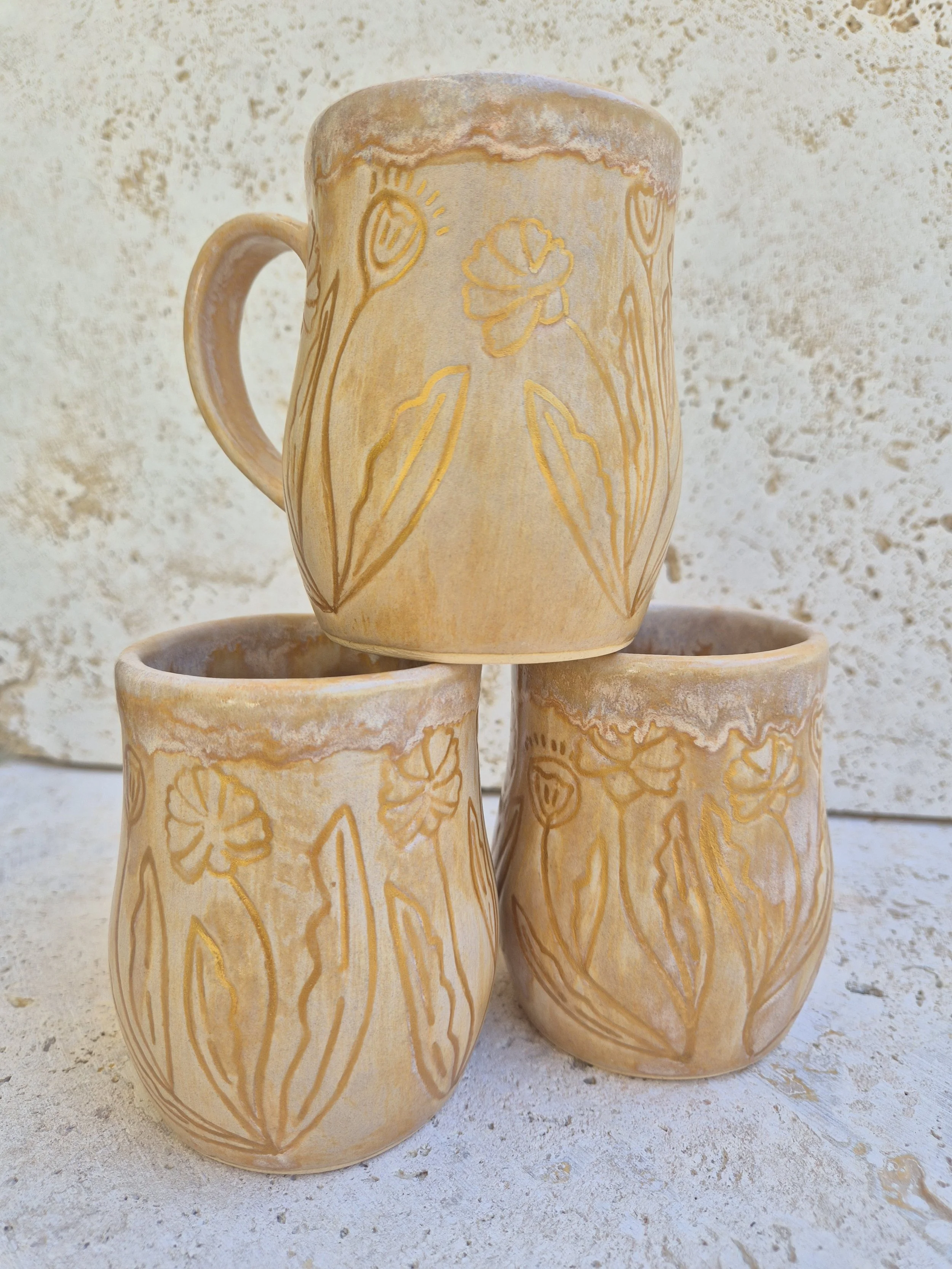 Three ceramic mugs with floral carvings in earthy tones, stacked with one on top of two, against a textured, light-colored background.