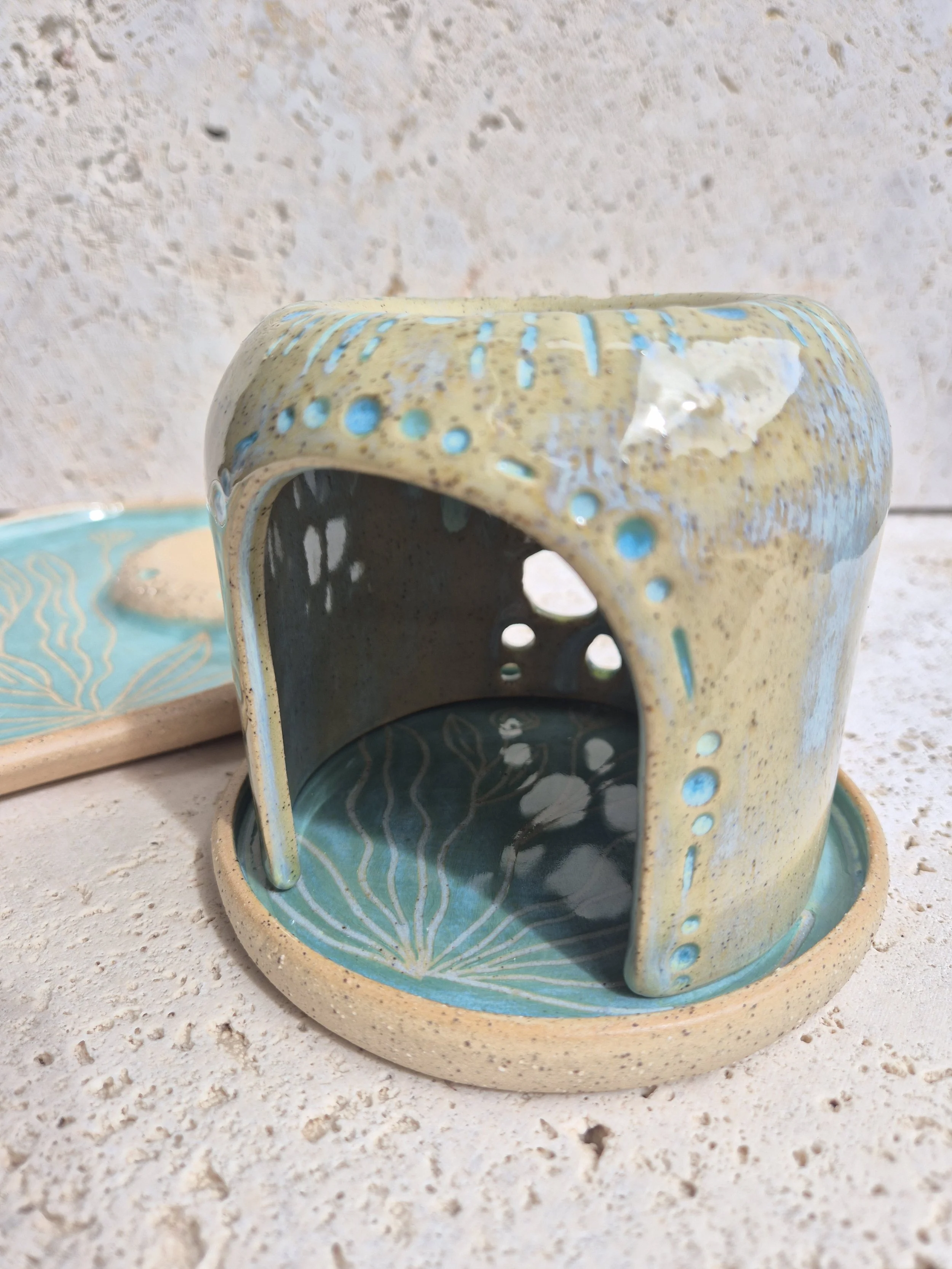 A ceramic incense holder with a rectangular shape and rounded edges, decorated with blue patterns on its beige surface, sits on a matching tray with a similar design, on a textured white surface.