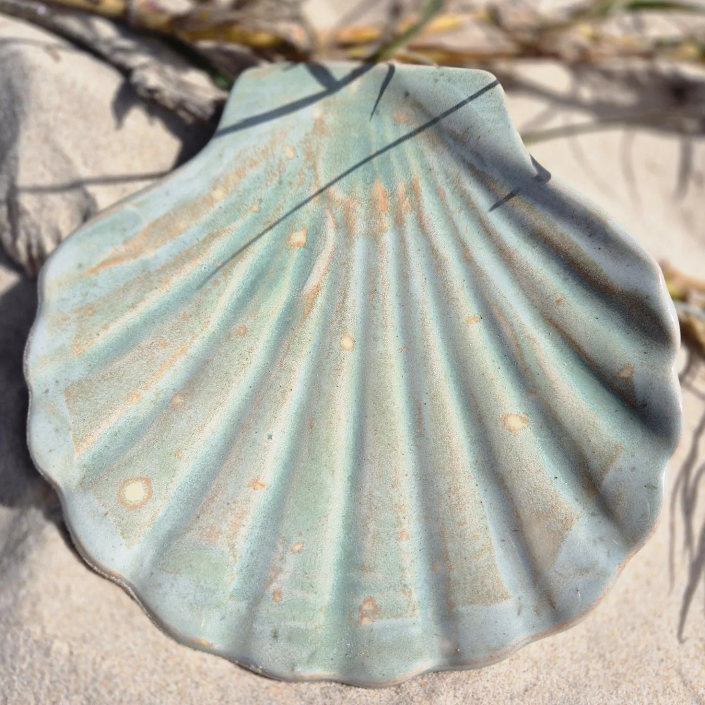 Close-up of a weathered, pale blue ceramic seashell-shaped dish on sandy ground with small rocks and dried twigs nearby.
