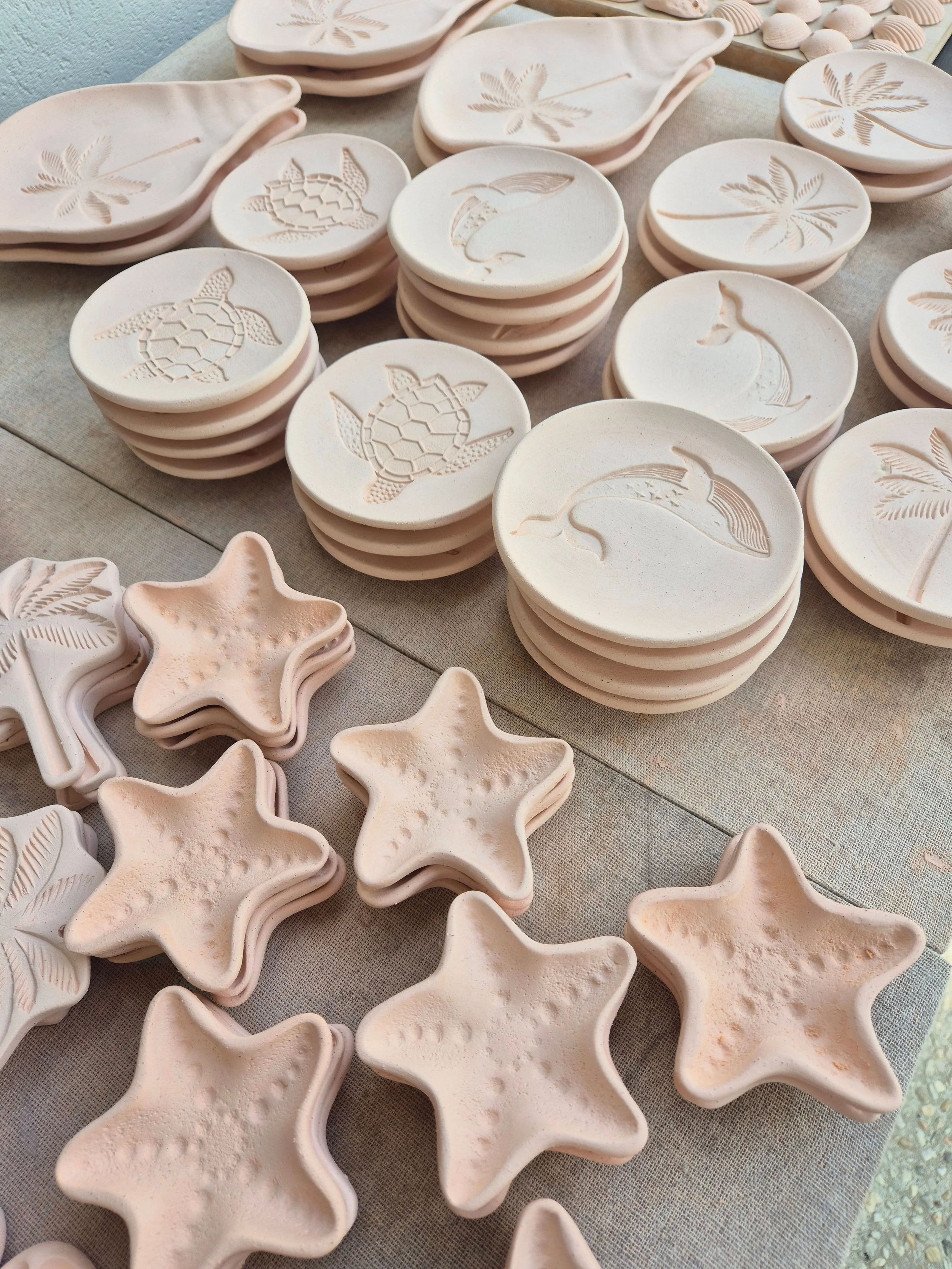 Stacks of white ceramic dishes and star-shaped pottery pieces featuring embossed and engraved turtle, dolphin, and palm tree designs, arranged on a beige fabric-covered surface.