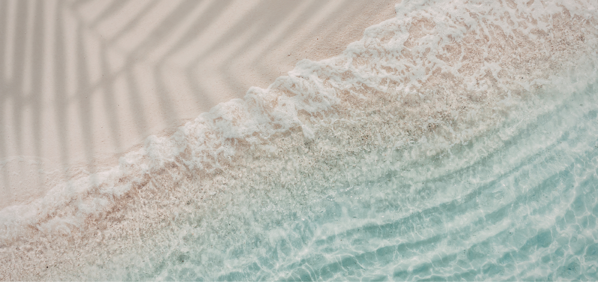 An aerial view of the shoreline where gentle waves meet light-colored sandy beach, creating white foam.