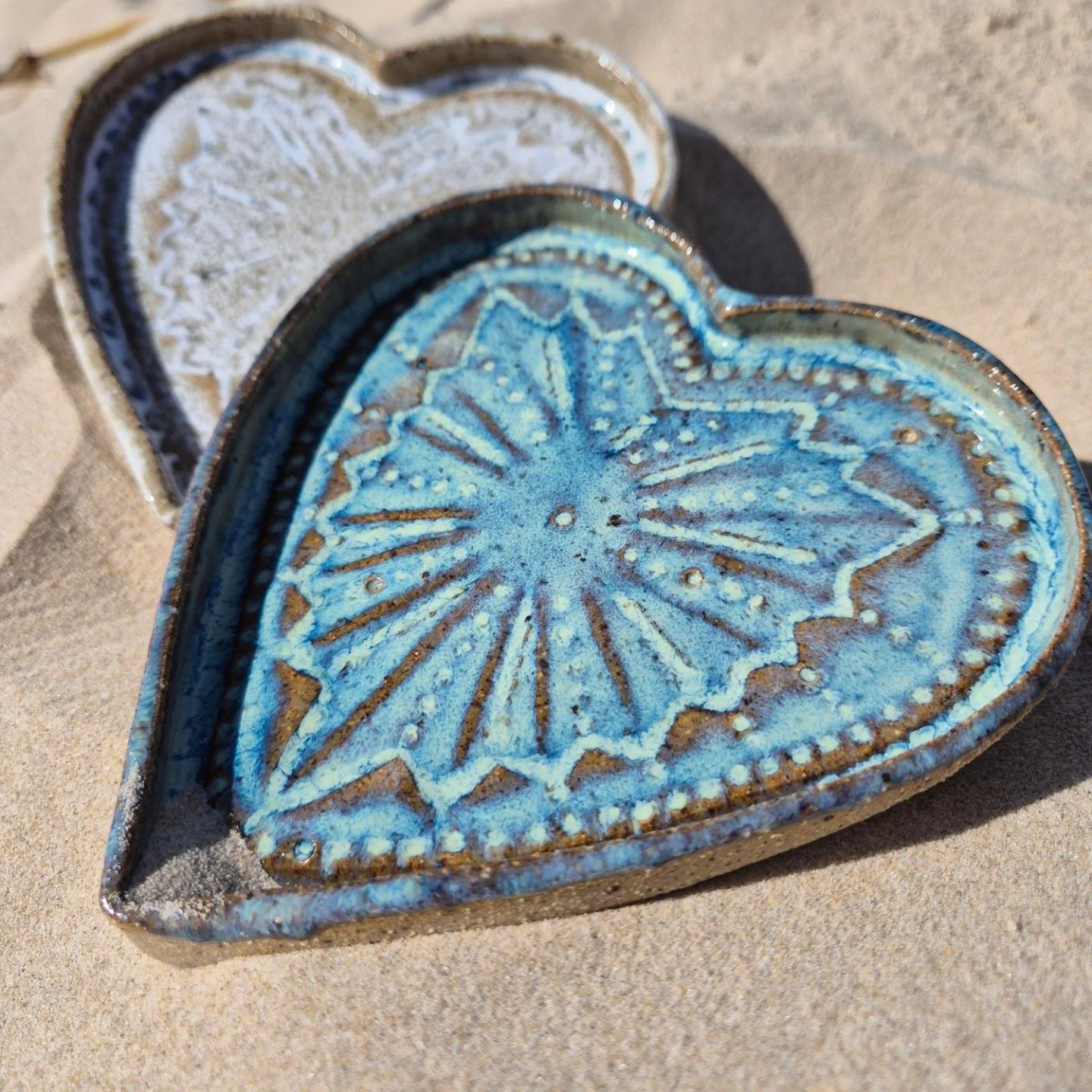 Two heart-shaped ceramic dishes with intricate designs, placed on sand. The foreground dish is blue with a starburst pattern, and the background dish is white with a similar pattern.