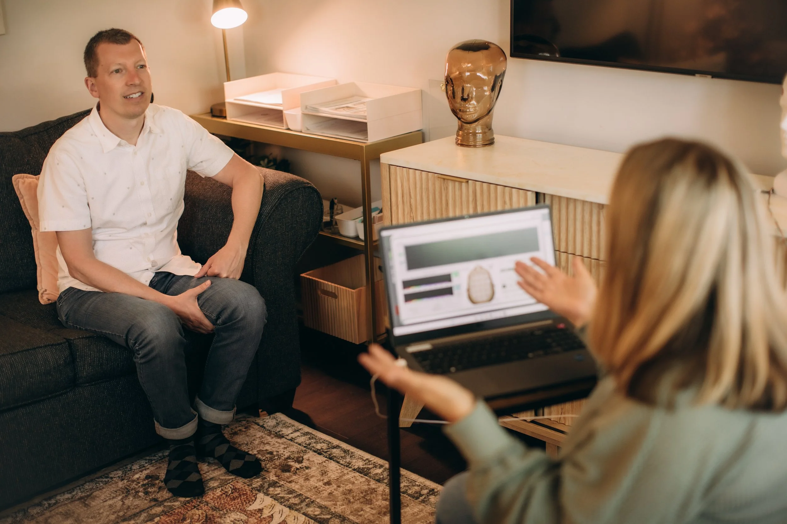 A man in a white shirt sitting on a dark sofa in a living room, talking to a person holding a laptop, with a lamp, decorative vase, and TV on the wall behind him.