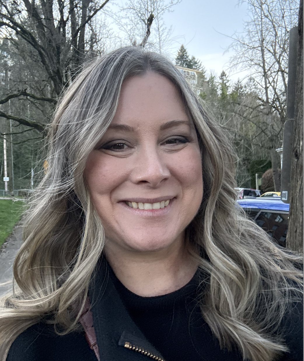 Smiling woman outdoors with trees and parked cars in the background.
