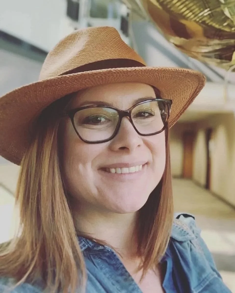 A woman smiling, wearing a wide-brimmed straw hat, glasses, and a denim shirt.