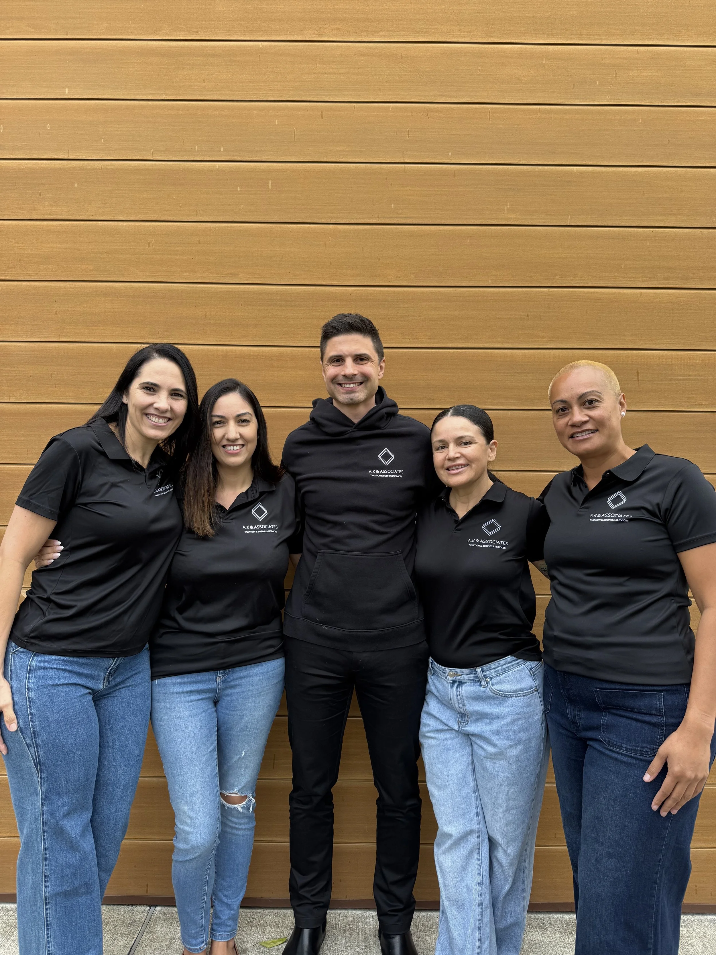 Group of five diverse people standing in front of a wooden wall, smiling, wearing black polo shirts with a logo and text that says 'A.K. & Associates'.