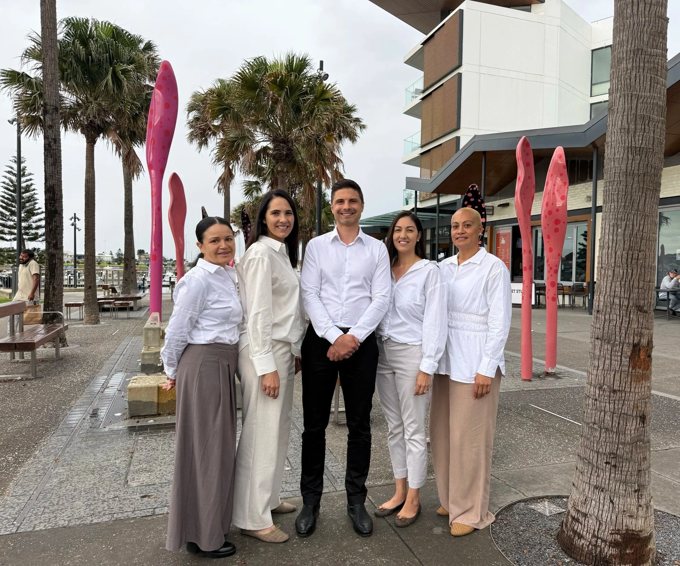 Group of five people standing outdoors near palm trees and a modern building, dressed in white and black, smiling at the camera.