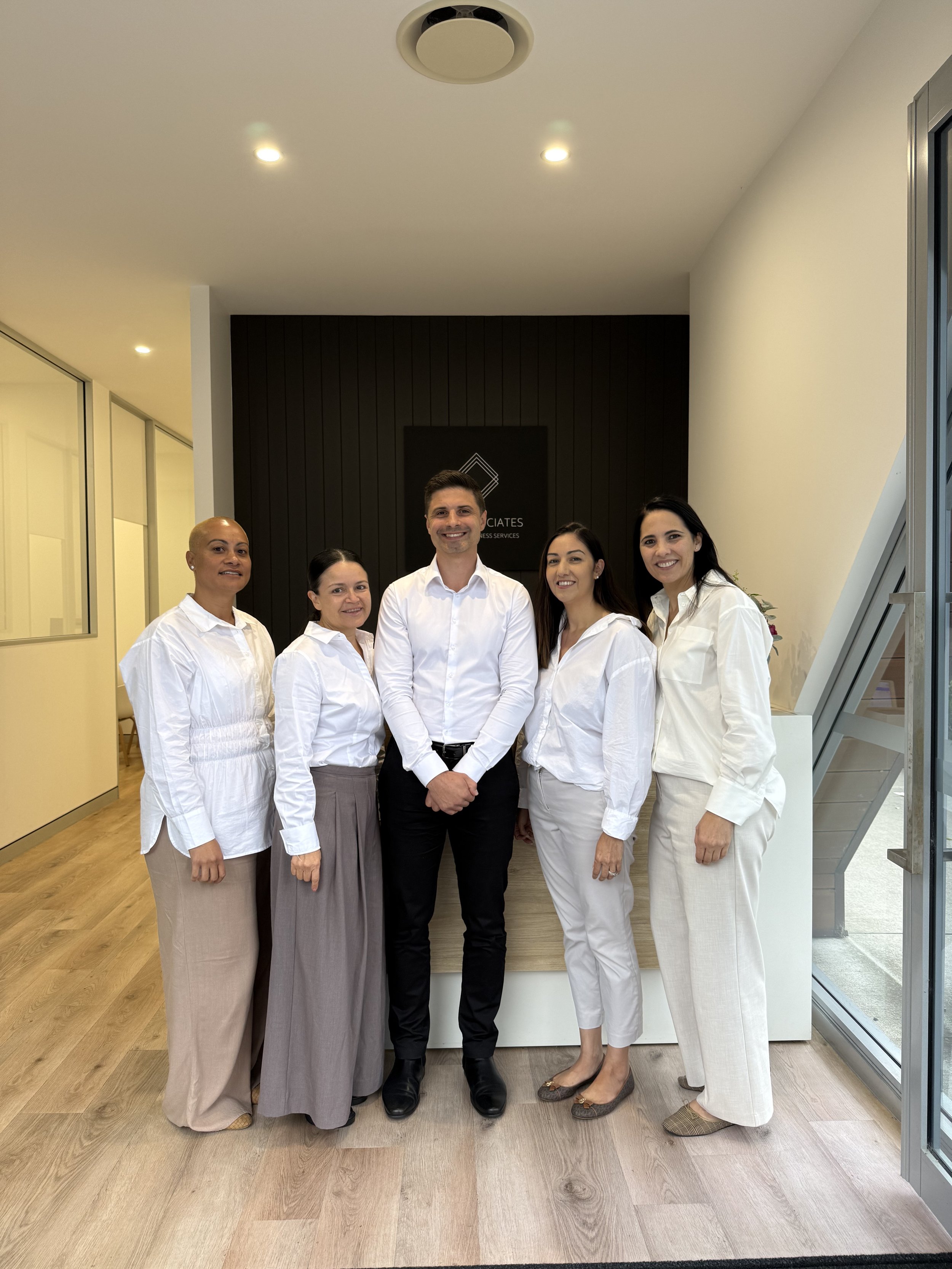 A diverse group of five professional women and one man standing together inside a modern office or lobby area, dressed in business casual attire, smiling at the camera.
