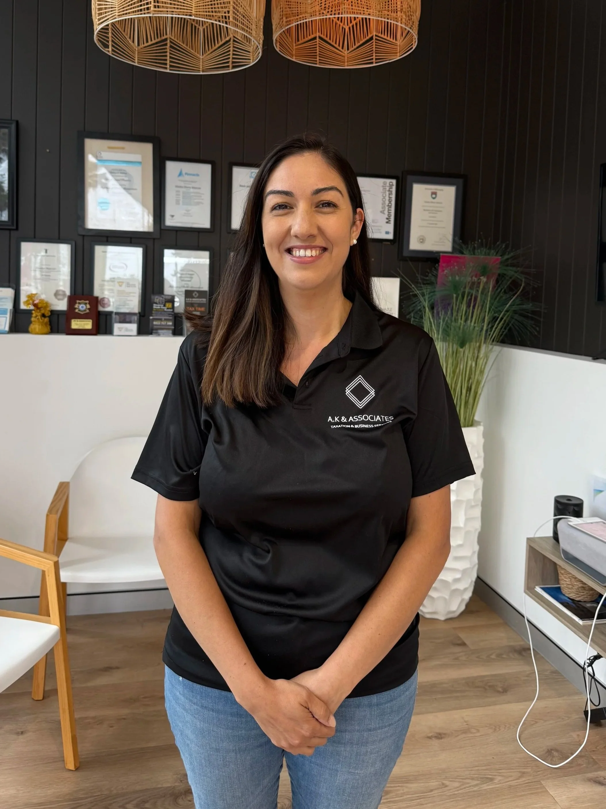 A woman with dark hair and pearl earrings smiling at the camera, standing in a room with framed certificates on a dark wall, a potted plant, and modern decor.