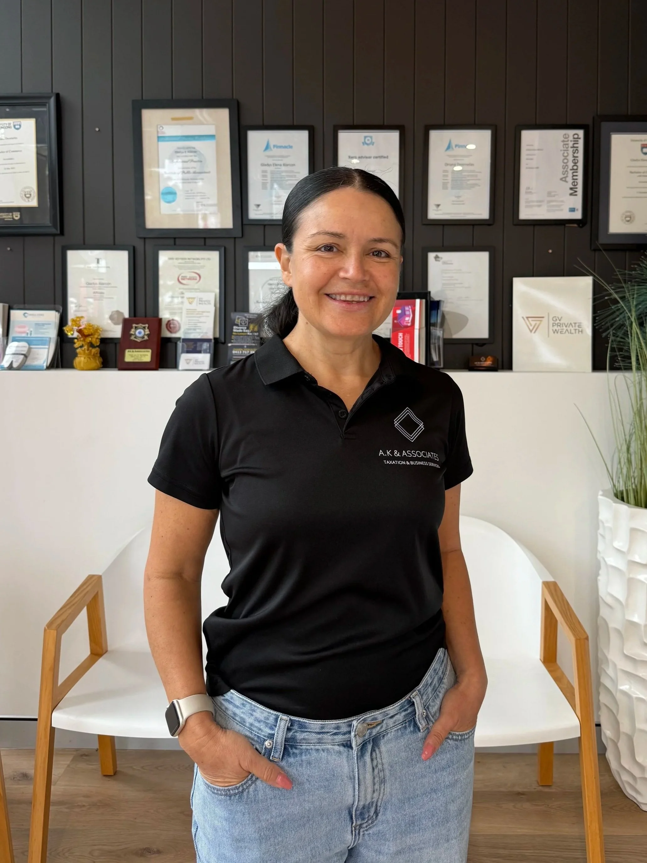 Smiling woman with dark hair and light skin standing indoors in front of framed certificates on a dark wooden-paneled wall, wearing a black polo shirt with logo and text, and light blue jeans, with white and silver smartwatch on her left wrist. She is standing between two white chairs with wooden arms.