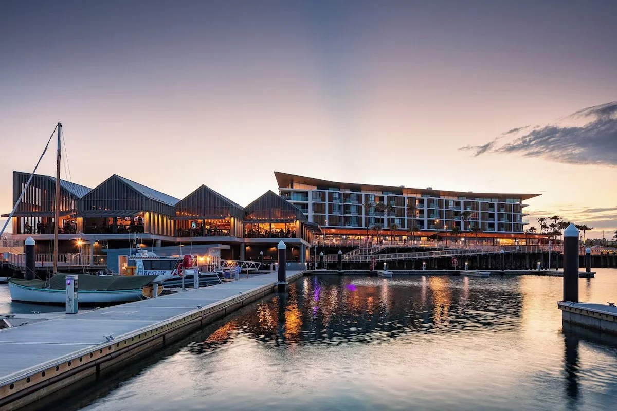 A modern waterfront restaurant and hotel with a marina during sunset, featuring boats docked at the pier and reflecting lights on the water.