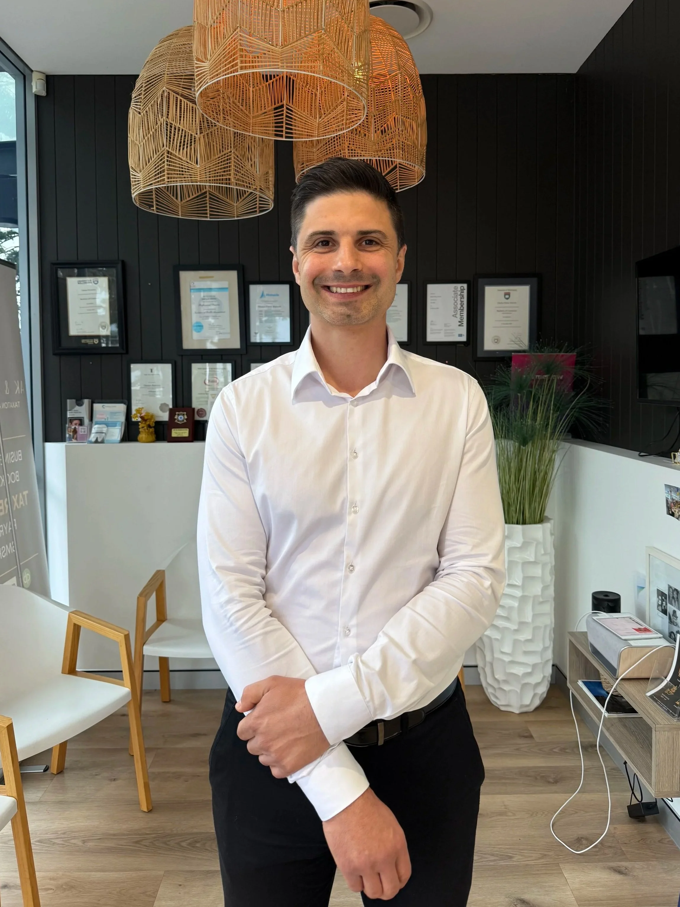 A smiling man wearing a white dress shirt and black pants stands in a modern office or waiting area, with framed certificates on a dark wall in the background, and wooden furniture including chairs and a small table.