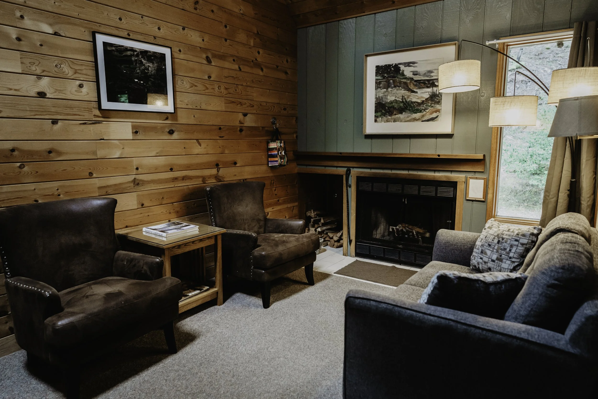 Cozy living room with wood-paneled walls, a fireplace, and a large window with curtains. Contains two brown leather armchairs, a gray sofa with patterned cushions, and a side table with magazines. Artwork hangs above the fireplace and on the wood-paneled wall.