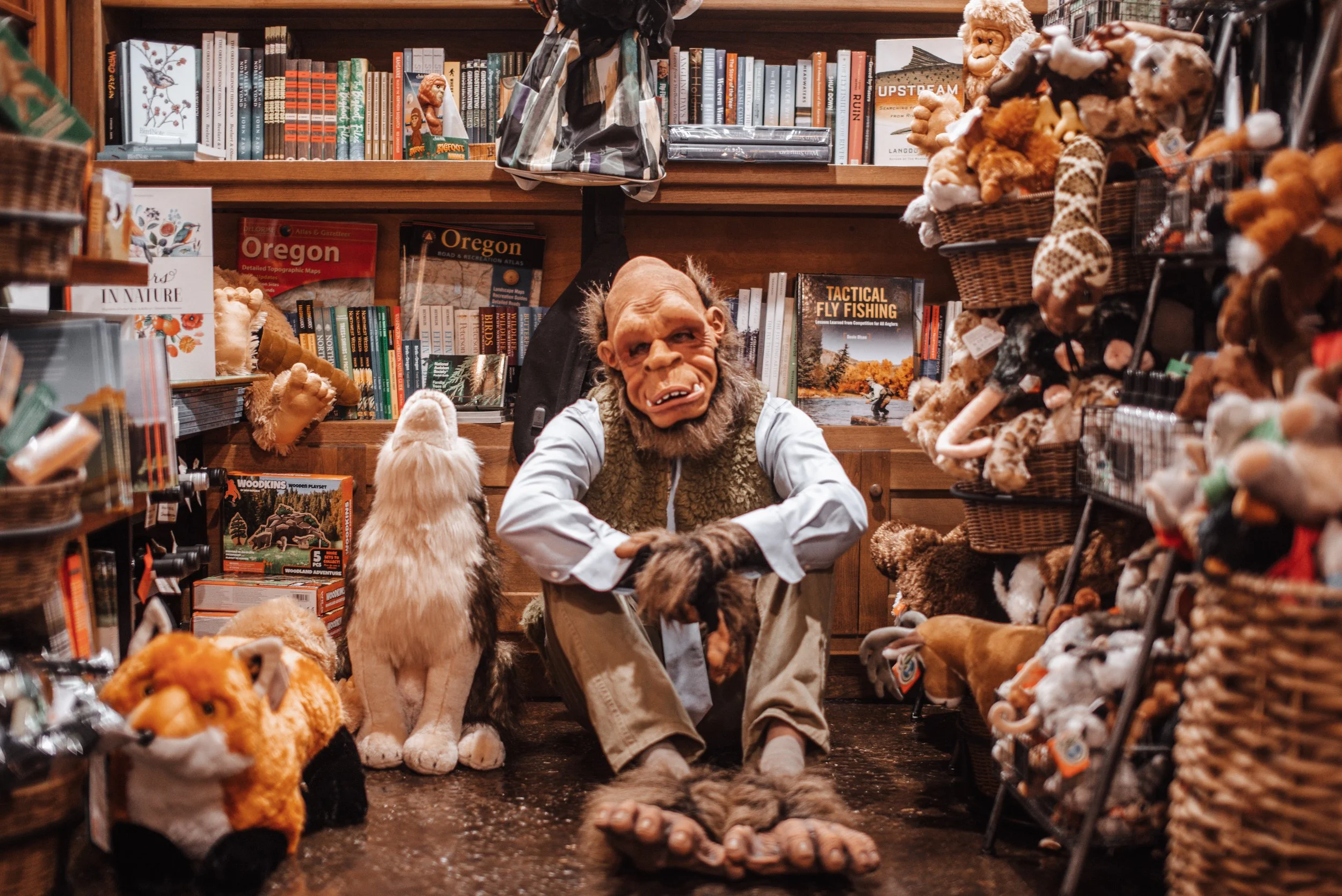 A person dressed in a Nosferatu costume with a mask, long gray hair, and a beard, sitting crossed-legged among shelves filled with plush animals, books, and stuffed toys in a store.