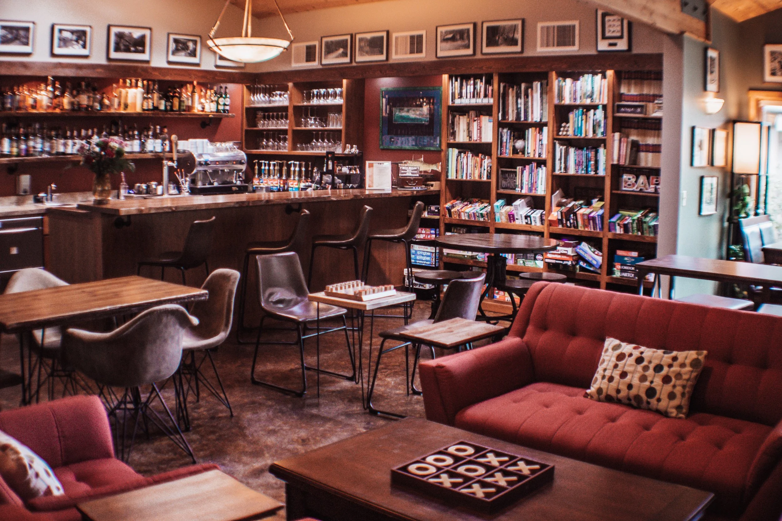 A cozy bar and lounge with a red couch, a wooden table with a game, several chairs, a bar counter with drinks, glasses, and a coffee machine, and bookshelves filled with books and framed pictures on the wall.
