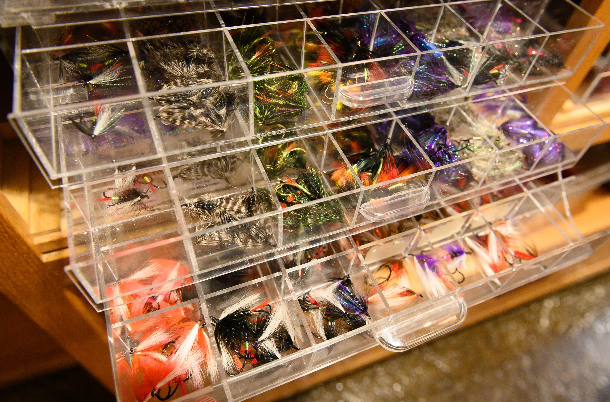Clear plastic storage box with multiple compartments filled with colorful fishing flies for fishing, arranged on a wooden surface.