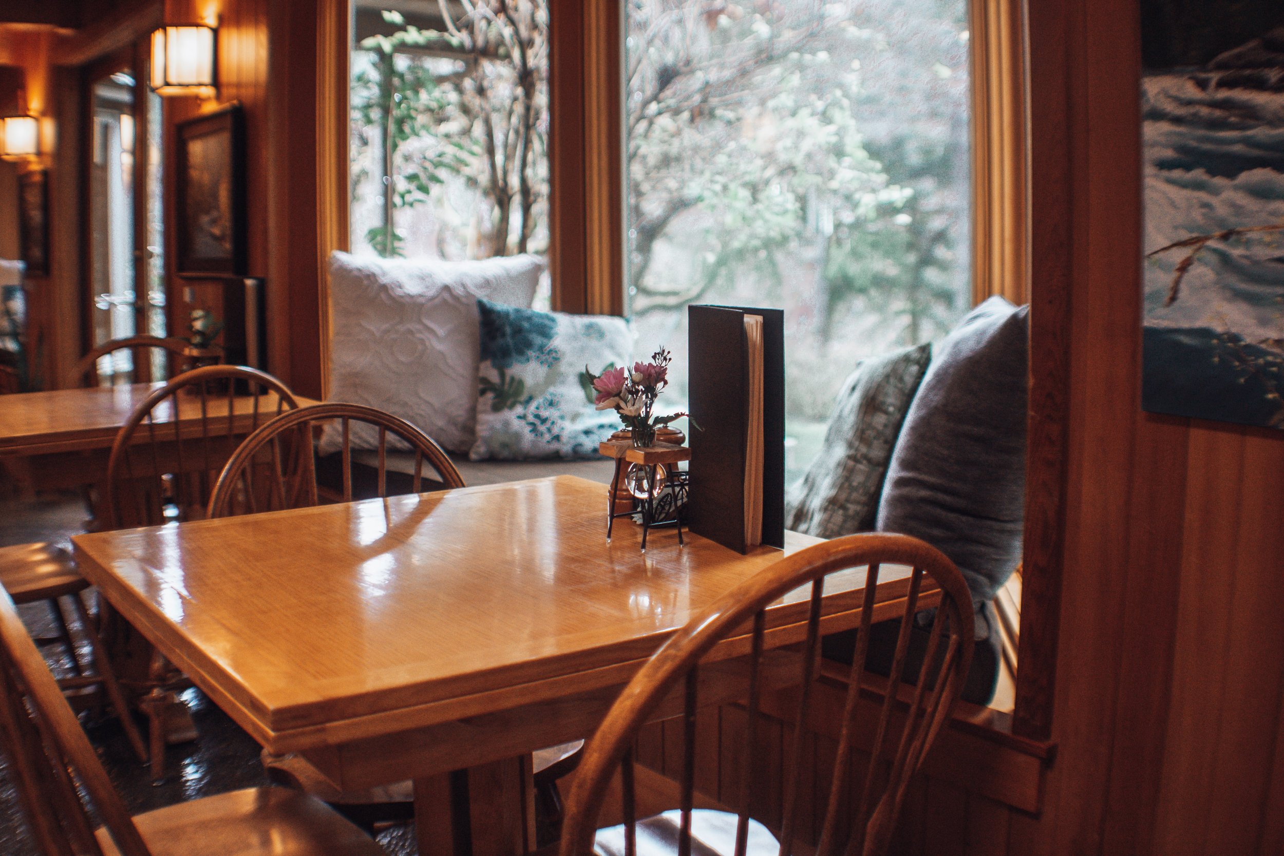 A cozy wooden dining area with a table and chairs, decorative pillows on window seats, and a view of trees outside through large windows.