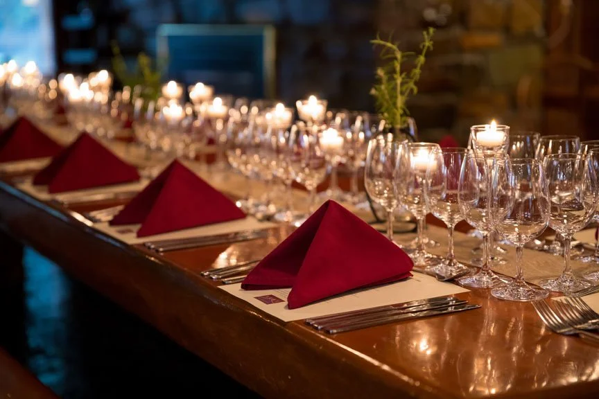 A long dining table set with red cloth napkins, glassware, and candles in a cozy, dimly lit environment.