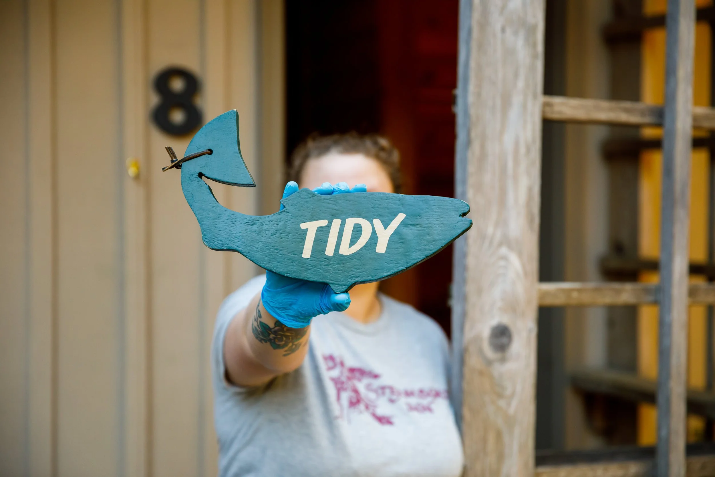 Person holding a blue wooden sign shaped like a fish with the word TIDY written on it, wearing blue gloves, in front of a wooden structure.