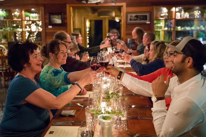 A group of people sitting around a long dining table raising glasses of red wine in a toast, enjoying a meal together in a cozy, warmly lit restaurant.