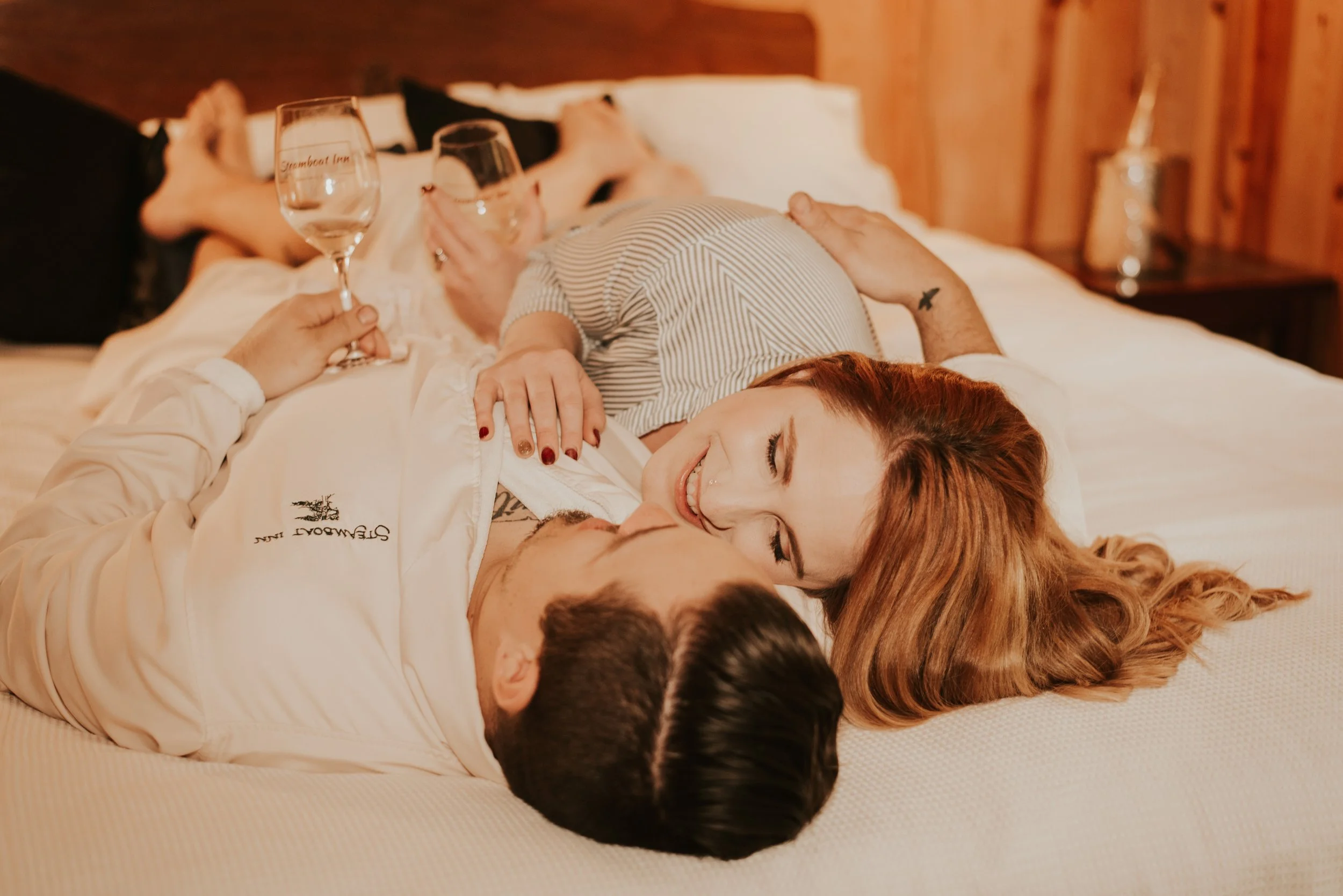 A couple lying on a bed, smiling and looking at each other, with wine glasses in hand, in a cozy wooden room.
