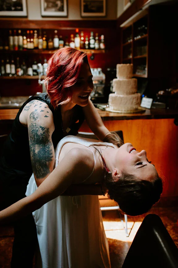Two women are sharing a moment at a bar, one with red hair and tattoos, smiling, leaning over a woman in a white dress who is lying back with her eyes closed.