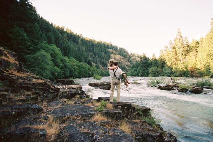 Couple standing on rocks by a river surrounded by trees and mountains, with one person holding a dog.