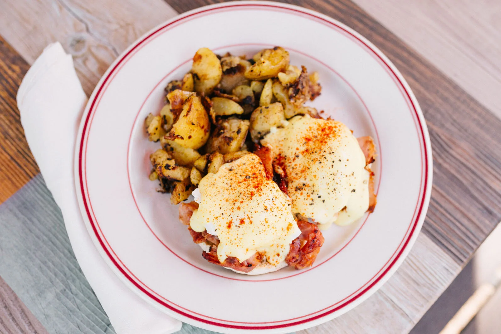 A white plate with a thin red stripe around the rim, containing eggs benedict topped with paprika and black pepper, served with roasted potatoes. The plate is on a wooden table with a white napkin underneath.