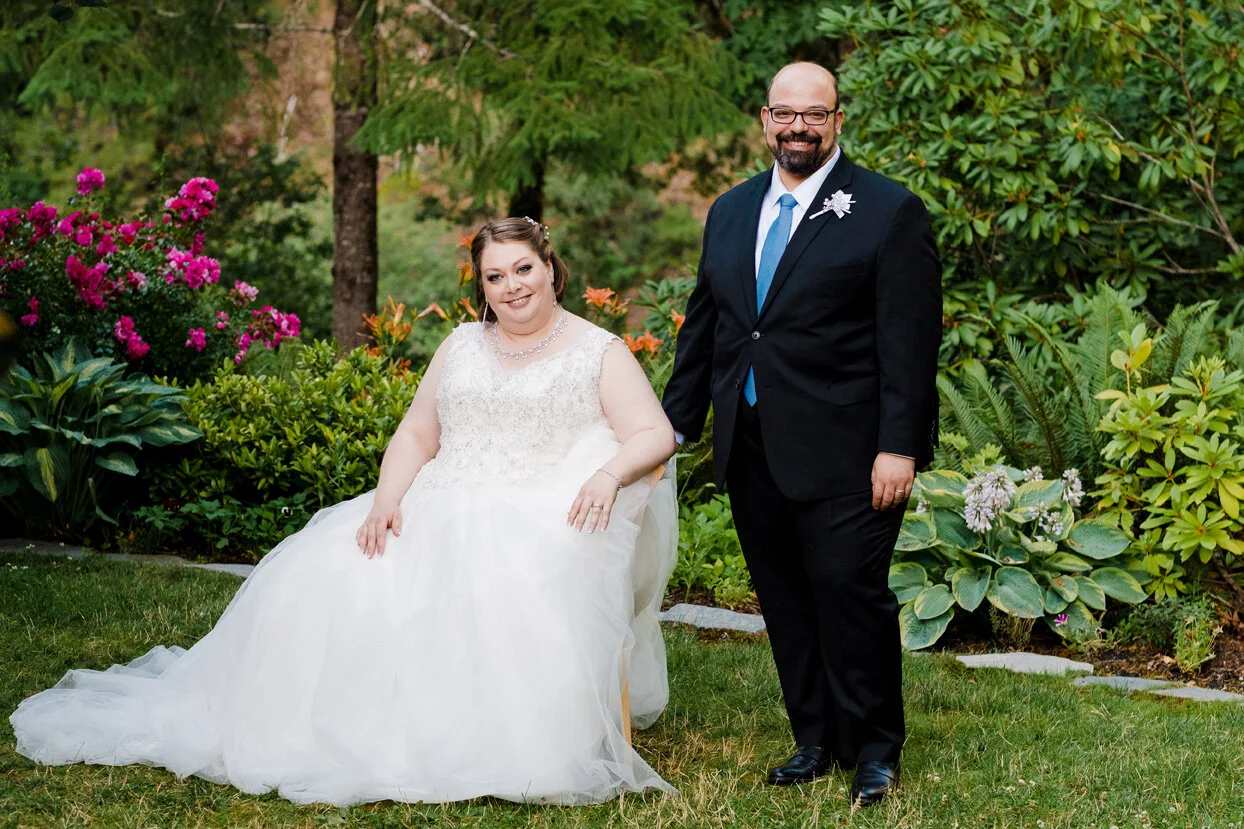 A bride and groom standing together outdoors in a garden surrounded by green plants and pink flowers. The bride is sitting on a chair, wearing a white wedding dress with lace and a pearl necklace. The groom is standing next to her, wearing a black su