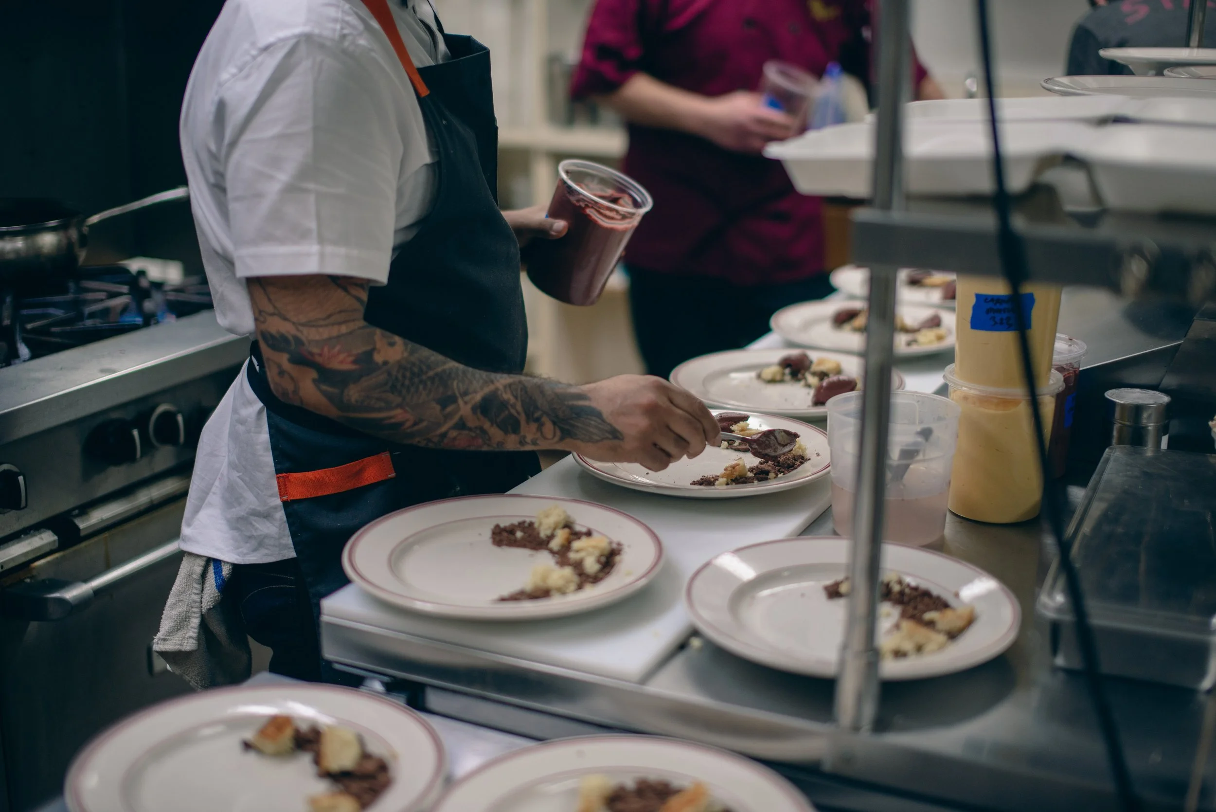 A chef with tattoos on his arm preparing plates of food in a busy kitchen, holding a cup of a dark beverage.