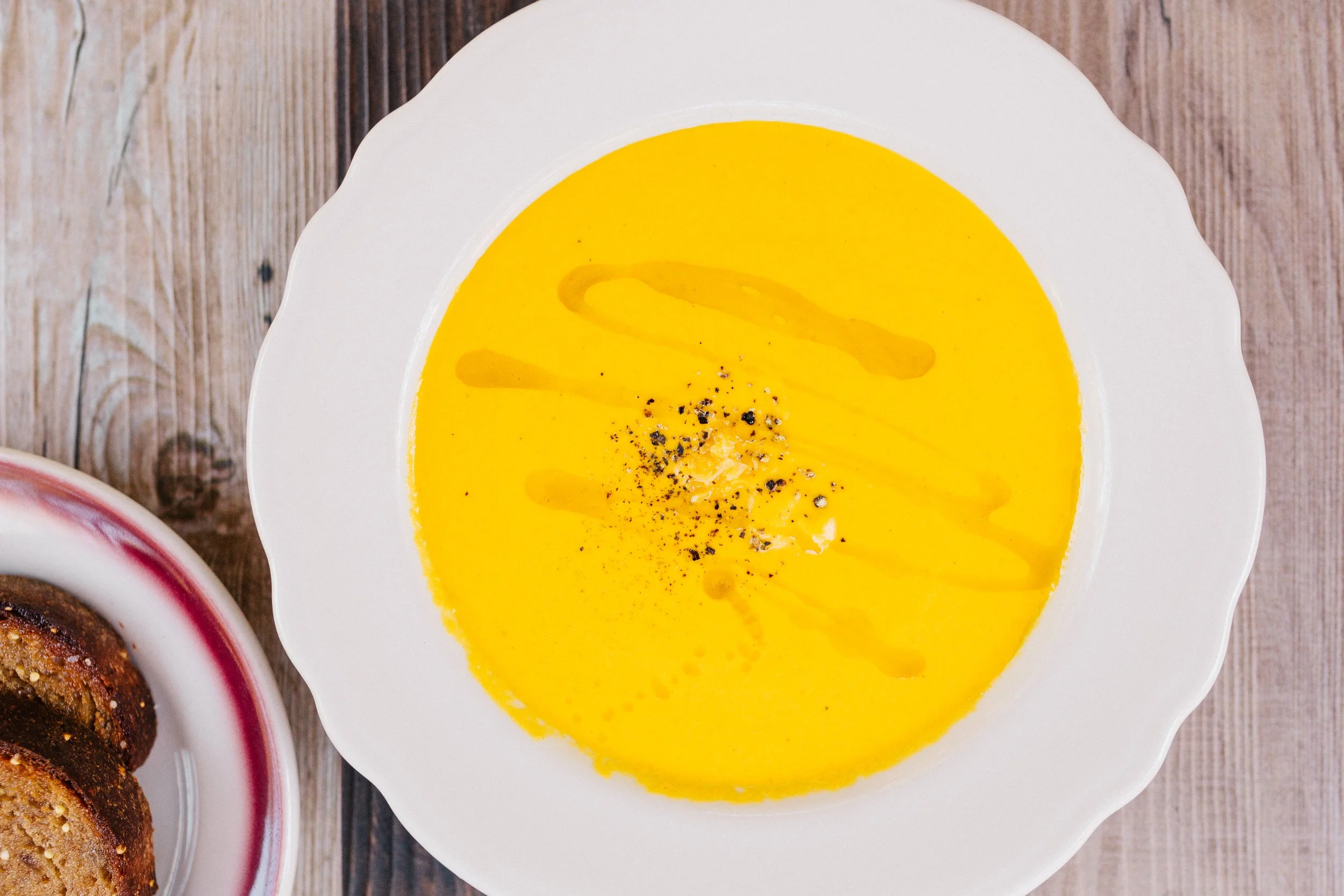A bowl of creamy yellow soup with a drizzle of oil, black pepper, and a swirl of cream, on a wooden surface.